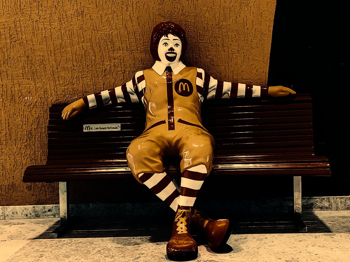 A clown sitting on a bench with his arms spread wide. Looks like he'd be the mascot of a famous fast food franchise.